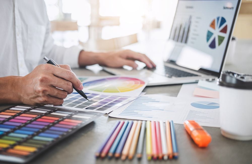 Personne travaillant sur des palettes de couleurs et des graphiques à l'aide d'un ordinateur portable, entourée de crayons de couleur et de documents graphiques.